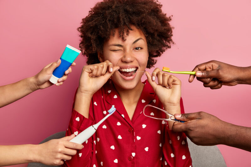 happy curly young woman brushes teeth with tooth floss cares about oral hygiene surrounded with toothpaste electric toothbrush tongue cleaner scaled 1