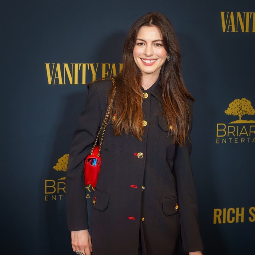 Anne Hathaway sorri em evento, de casaco preto com botões dourados e bolsa vermelha
