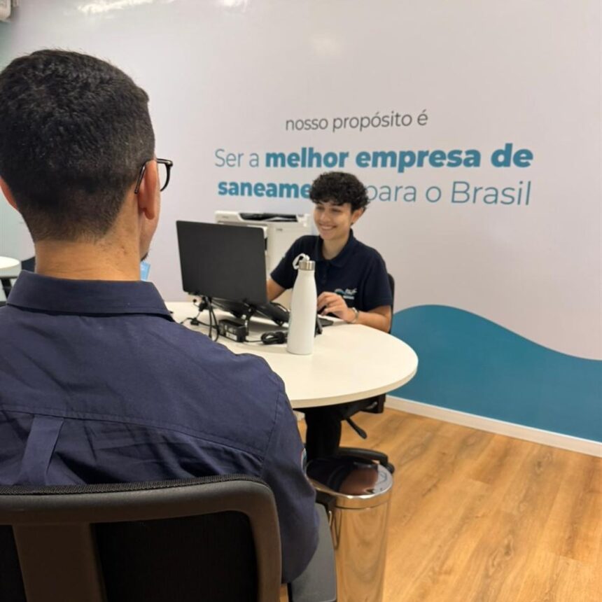Cliente de costas, sentado diante de uma atendente sorridente em mesa redonda com computador, em loja com parede azul e branca da Iguá