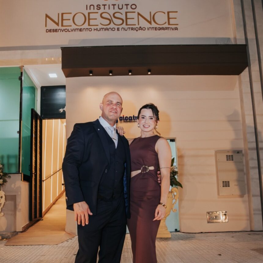Homem de terno escuro e mulher de vestido vinho posam abraçados na entrada do Instituto NeoEssence, à noite