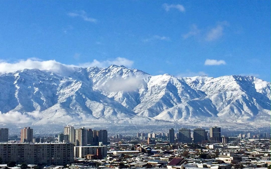 Vista da cidade de Santiago, no Chile, com prédios em primeiro plano e a Cordilheira dos Andes coberta de neve ao fundo, sob céu azul